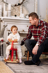 Obraz na płótnie Canvas Smiling father with little cute child in warm plush costume sits and swinging on the rocking wooden red deer chair in a room decorated with Christmas garlands and lights. Magic moment with family.