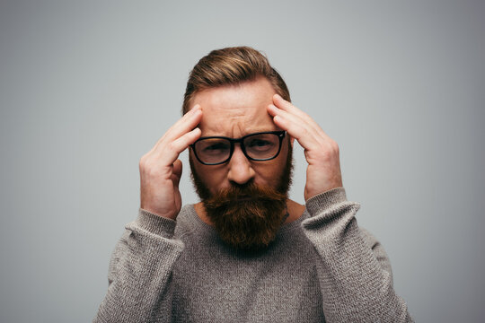 Stressed man in eyeglasses looking at camera while touching forehead isolated on grey.