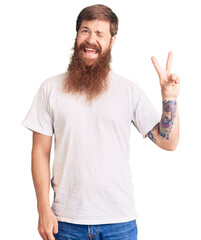 Handsome young red head man with long beard wearing casual white tshirt smiling with happy face winking at the camera doing victory sign. number two.
