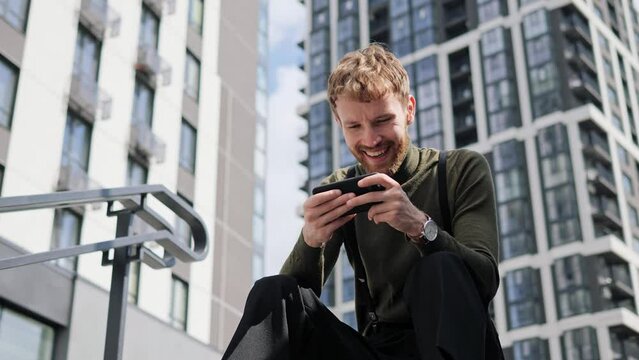 Excited cool guy gamer male urban professional businessman using mobile cell phone app sitting on the stairs in city play online video games enjoying free time relaxing using smartphone outdoor. 