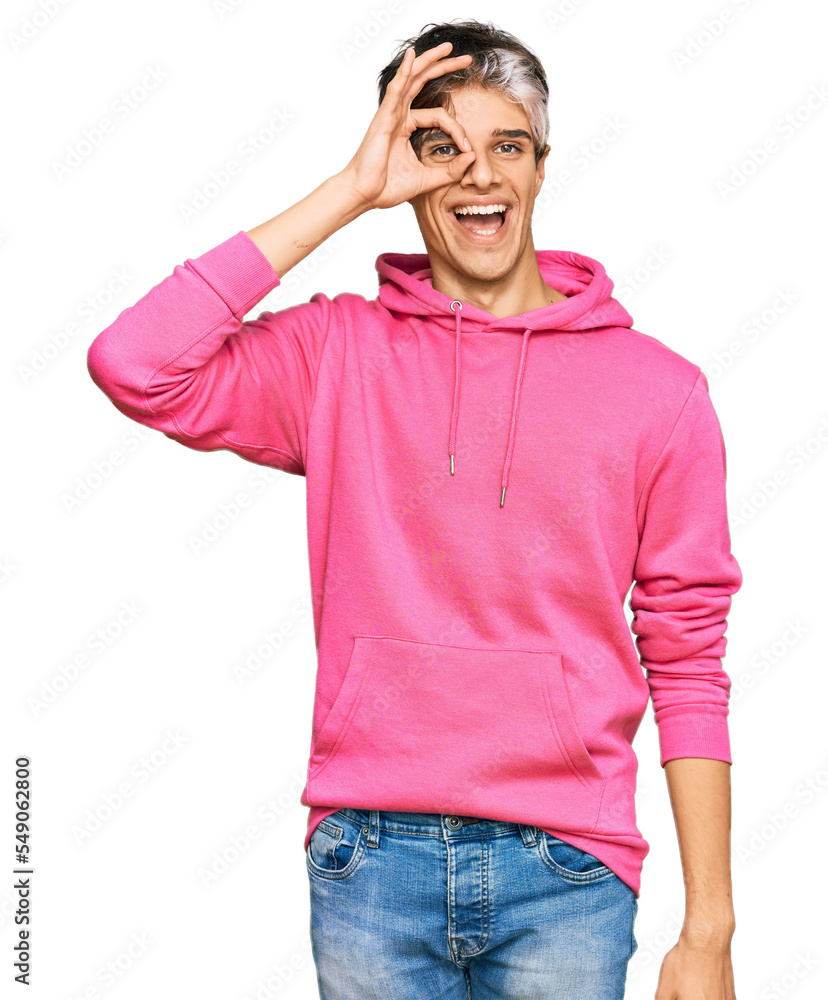 Sticker Young hispanic man wearing casual pink sweatshirt doing ok gesture with hand smiling, eye looking through fingers with happy face.