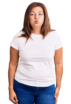 Middle age latin woman wearing casual white tshirt puffing cheeks with funny face. mouth inflated with air, crazy expression.