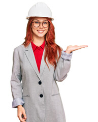 Young redhead woman wearing architect hardhat smiling cheerful presenting and pointing with palm of hand looking at the camera.
