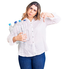Young caucasian woman holding recycling plastic bottles with angry face, negative sign showing dislike with thumbs down, rejection concept