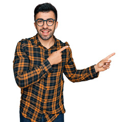 Hispanic man with beard wearing casual clothes and glasses smiling and looking at the camera pointing with two hands and fingers to the side.