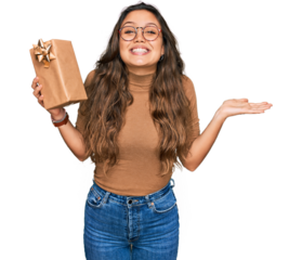 Young hispanic girl holding gift celebrating victory with happy smile and winner expression with raised hands