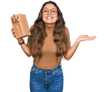 Young hispanic girl holding gift celebrating victory with happy smile and winner expression with raised hands