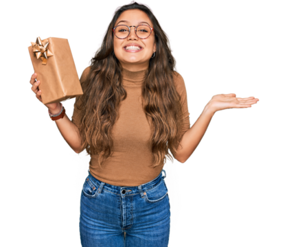 Young hispanic girl holding gift celebrating victory with happy smile and winner expression with raised hands