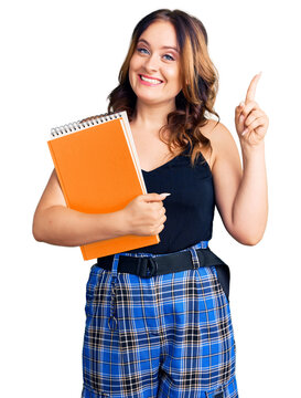Young beautiful caucasian woman holding book surprised with an idea or question pointing finger with happy face, number one