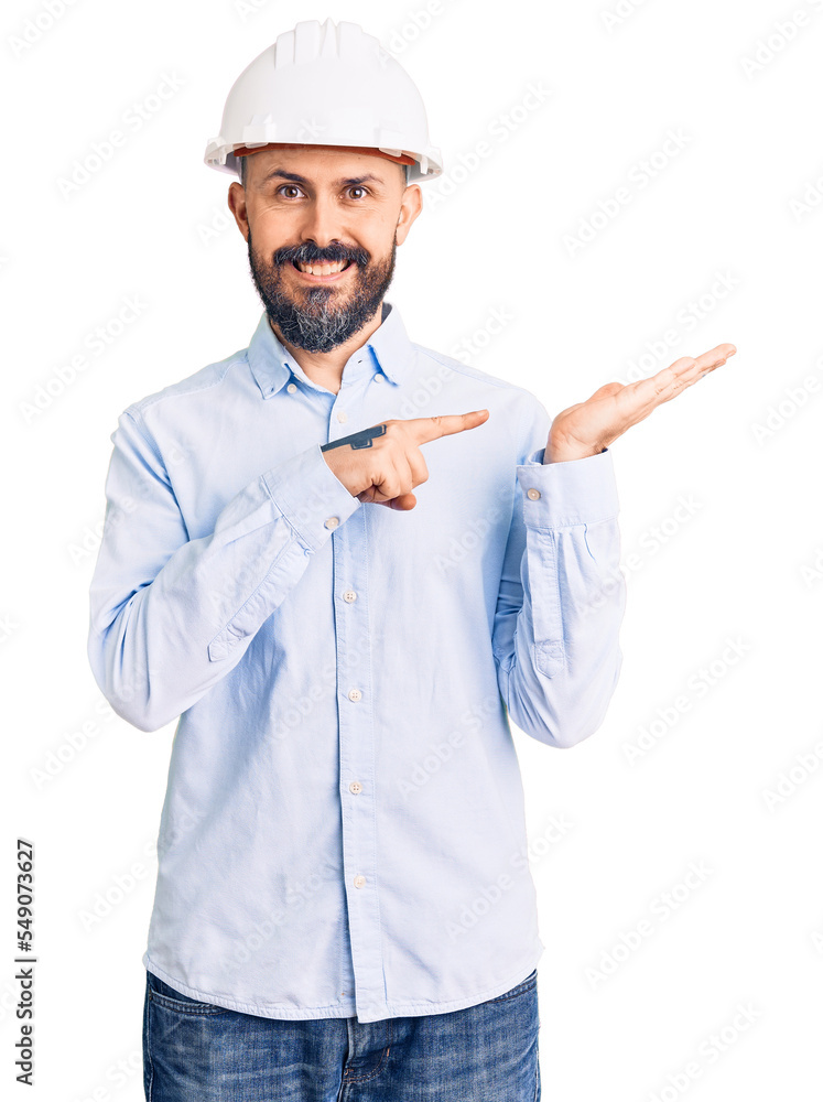 Poster young handsome man wearing architect hardhat amazed and smiling to the camera while presenting with 