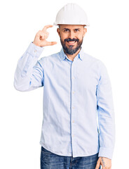 Young handsome man wearing architect hardhat smiling and confident gesturing with hand doing small size sign with fingers looking and the camera. measure concept.