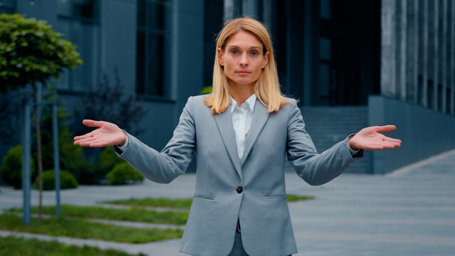 Confused serious pensive puzzled businesswoman standing outdoors spreads arms shrugs unsure caucasian woman doesn't know answer looks at camera showing ignorance gesture sign doubt shrugging shoulders
