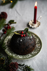 Traditional Christmas pudding decorated with holly on vintage plate, lit candle and natural decorations on linen tablecloth with Christmas lights on background.