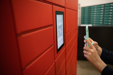 A woman scans a red code to pick up a parcel at a parcel machine. Automated Postal Box.