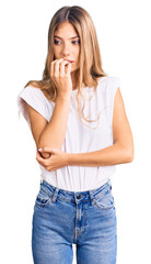 Beautiful caucasian woman with blonde hair wearing casual white tshirt looking stressed and nervous with hands on mouth biting nails. anxiety problem.