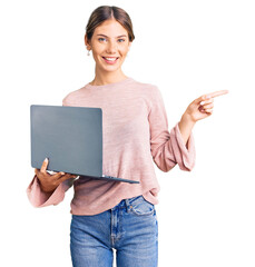 Beautiful caucasian woman with blonde hair working using computer laptop smiling happy pointing with hand and finger to the side