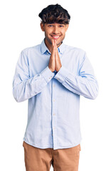 Young hispanic man wearing casual shirt praying with hands together asking for forgiveness smiling confident.