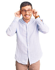 Handsome young man with bear wearing elegant business shirt and glasses smiling pulling ears with fingers, funny gesture. audition problem