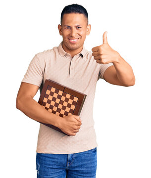 Young handsome latin man holding chess smiling happy and positive, thumb up doing excellent and approval sign