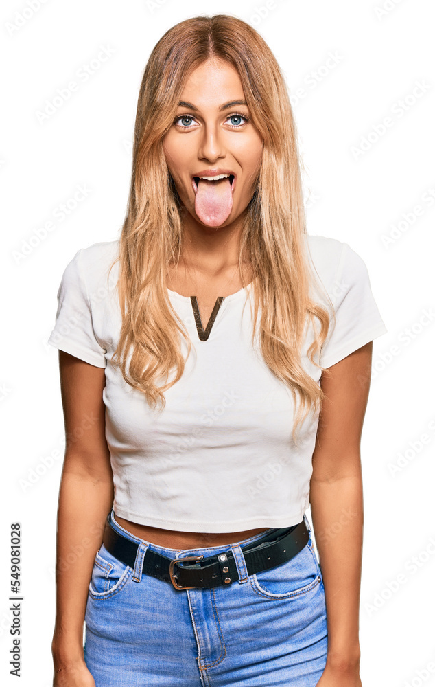 Poster beautiful blonde young woman wearing casual white tshirt sticking tongue out happy with funny expres