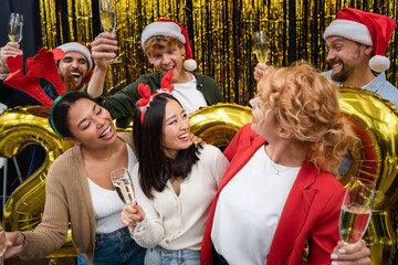Fototapeta premium Smiling multiethnic businesswomen holding champagne near balloons and businessmen during Christmas party in office