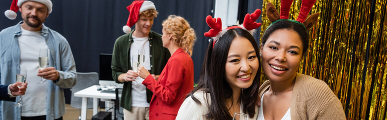Asian and multiracial businesswomen in Christmas headbands looking at camera near tinsel in office, banner