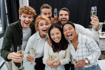 Cheerful interracial business people holding glasses of champagne and looking at camera in office