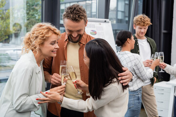 Smiling businessman hugging interracial colleagues with champagne in office