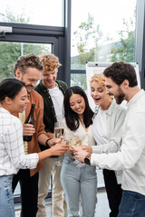 Fototapeta na wymiar Happy multiethnic business people holding glasses near colleague with bottle of champagne in office