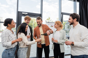 cheerful man pouring champagne near multicultural friends during corporate party