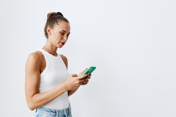 A young woman with beautiful and tanned skin looks at the phone screen in her hands wearing a white T-shirt and blue jeans against a white wall