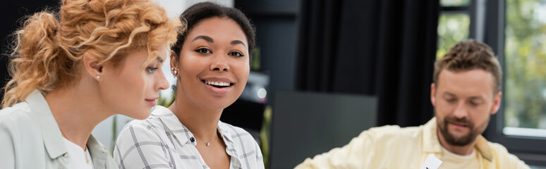 happy multiracial woman looking at camera near colleagues working in office, banner