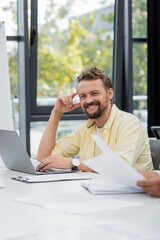 positive bearded businessman smiling at camera near laptop at workplace