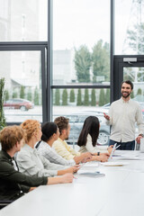 Fototapeta premium positive bearded businessman pointing with hand while talking to multiethnic colleagues