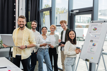 cheerful multiethnic corporate team looking at camera near flip chart with infographics