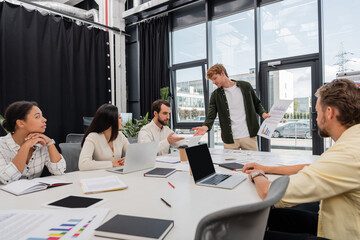young businessman with charts talking to colleague pointing at digital tablet during meeting with interracial team