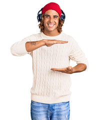Young hispanic man listening to music using headphones gesturing with hands showing big and large size sign, measure symbol. smiling looking at the camera. measuring concept.