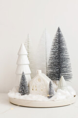 Modern christmas scene, miniature snowy village on white table. Merry Christmas! Stylish little Christmas trees and house. Winter hygge, scandinavian  monochromatic decor.