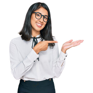 Beautiful asian young woman wearing business shirt and glasses amazed and smiling to the camera while presenting with hand and pointing with finger.