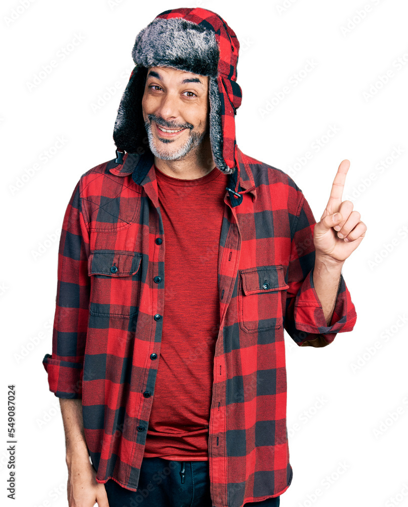 Poster Middle age hispanic man with grey hair wearing fluffy earmuff hat showing and pointing up with finger number one while smiling confident and happy.