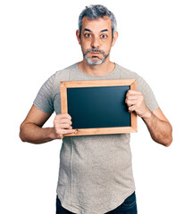 Middle age hispanic man with grey hair holding blackboard puffing cheeks with funny face. mouth inflated with air, catching air.