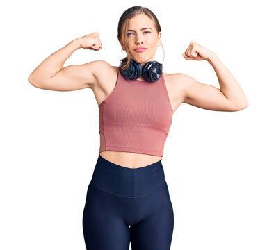 Beautiful caucasian young woman wearing gym clothes and using headphones showing arms muscles smiling proud. fitness concept.