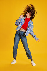 Portrait of young emotional girl posing in stylish casual clothes, singing in lollipop isolated over yellow background