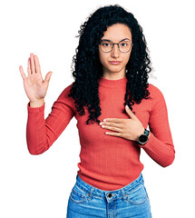 Young hispanic woman with curly hair wearing glasses swearing with hand on chest and open palm,...