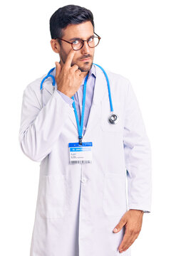 Young hispanic man wearing doctor uniform and stethoscope pointing to the eye watching you gesture, suspicious expression