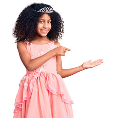 African american child with curly hair wearing princess crown amazed and smiling to the camera while presenting with hand and pointing with finger.