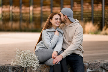 Cheerful young woman and man sitting outdoor. Guy kisses girlfriend. Love story