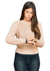 Young brunette woman wearing casual winter sweater checking the time on wrist watch, relaxed and...