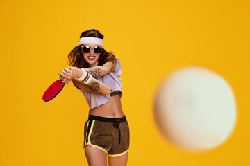 Portrait of young emotive stylish girl posing, playing table tennis isolated over yellow...