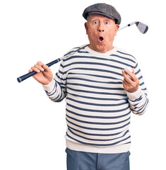 Senior handsome grey-haired man holding golf club and ball scared and amazed with open mouth for...